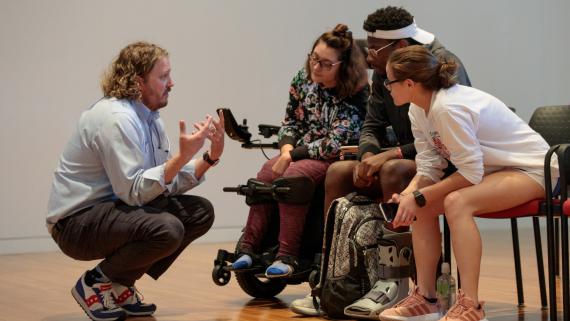 Photo of college students sitting and a professor crouching, having a discussion.