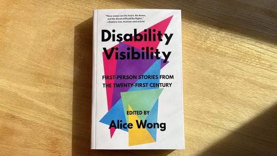 Photo of the book cover, with black text, multi-colored triangles, and off-white background. The cover is displayed on a wooden table.